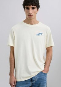 T-shirt a maniche corte color crema con una grafica blu sul lato sinistro del petto. Fabbricato in tessuto morbido con una vestibilità rilassata.
