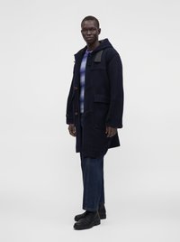 Mann trägt einen dunkelblauen Kapuzenmantel über einem blau-weiß gestreiften Pullover, dunkle Jeans und schwarze Schnürstiefel, steht und blickt nach vorne.