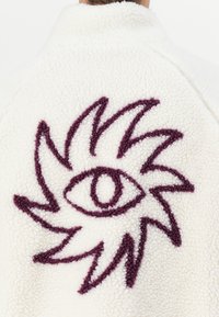 White fleece jacket featuring a textured burgundy embroidered eye design with spiky accents on the back. Soft and cozy material.