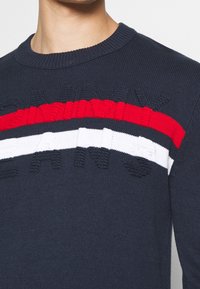 Pull en tricot marine avec des rayures horizontales rouges et blanches ; présente des lettres texturées. Col rond et poignets côtelés, texture douce, design décontracté.