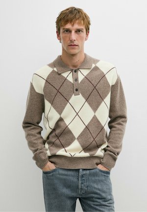Jeune homme aux cheveux châtains clairs portant un pull beige et blanc à motifs argyle et un jean bleu, les mains dans les poches, debout devant un fond blanc uni.