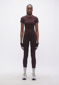 Γυναίκα όρθια, ντυμένη με σκούρα καφέ αθλητική περιβολή, λευκές κάλτσες, λευκά αθλητικά παπούτσια και μαύρα-άσπρα γάντια προπόνησης.