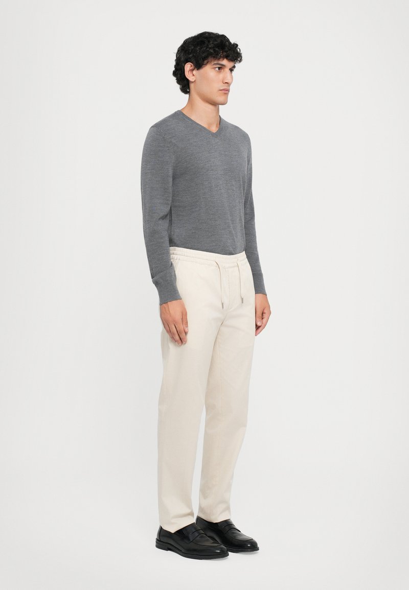 Grijze lange mouw v-hals trui, crèmekleurige broek met elastische taille, zwarte schoenen. Model staat zijwaarts tegen een effen witte achtergrond.