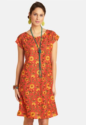 Woman wearing a knee-length orange dress with yellow floral patterns, paired with green beaded necklace, green earrings, and a multicolored bracelet.