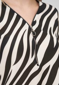 Zebra-patterned top in cream and black, featuring a front zipper with a circular pull. Textured fabric with dynamic stripe design.