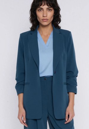 Kobieta o ciemnych, kręconych włosach, ubrana w turkusową marynarkę i spodnie do kompletu, na jasnoniebieskiej bluzce z dekoltem w serek, stoi na jednolitym tle.