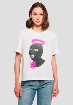 Junge Frau mit schulterlangem braunem Haar trägt weißes T-Shirt mit schwarzer Balaklava-Gesichtsmaske mit rosa Heiligenschein und Graffiti-Text, blaue Jeans.