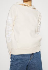 Persona che indossa un felpa crema di Calvin Klein Jeans con tasca frontale e testo bianco del logo sul petto e sulle maniche, abbinata a jeans blu.