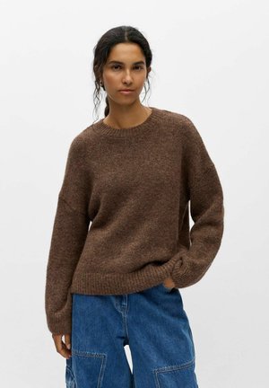 Jeune femme aux cheveux foncés portant un pull en maille marron et un jean bleu ample, debout avec une main dans la poche devant un fond blanc uni.
