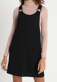 Czarna sukienka na szelkach z denimu z dwoma przednimi kieszeniami, okrągłymi ozdobami z metalu oraz regulowanymi szelkami, noszona na jasnoróżowej bluzce z długim rękawem.