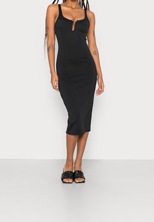 Robe noire ajustée avec un décolleté en trou de serrure, des bretelles larges et une longueur midi. Tissu lisse avec des coutures de contour. Associée à des sandales noires tissées à enfiler.