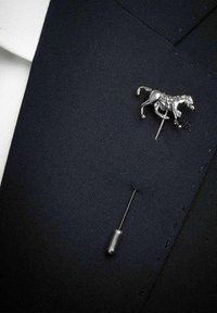 Silver hästformad slipsnål fäst vid en mörk tyglik kostym, åtföljd av en silversäkerhetsnål. Texturerad marinblå tygbakgrund.