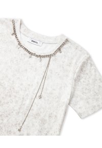 T-shirt gris clair avec un motif texturé, comportant un détail en chaîne argentée autour de l'encolure avec de petits pendentifs décoratifs.