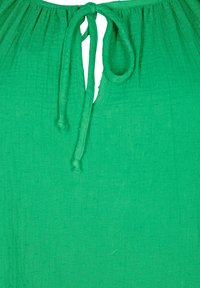 Maglietta verde caratterizzata da un tessuto testurizzato con un sottile motivo a pois. Presenta un dettaglio a chiave sul collo e una chiusura a nodo all'altezza del collo.