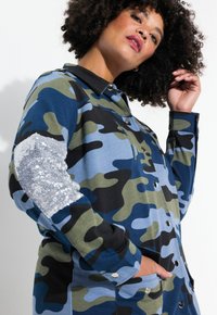 Chaqueta de camuflaje en azul y verde, con acentos en negro y un parche de lentejuelas en la manga. Incluye botones y bolsillos.