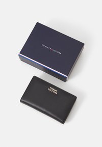 Black leather wallet with gold logo, placed next to a navy blue box with white and red stripes and "TOMMY HILFIGER" branding on top.