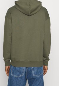 Sudadera con capucha color verde oliva, de corte holgado y puños acanalados, combinada con vaqueros de mezclilla azul. Tejido liso, diseño sencillo, sin logos.