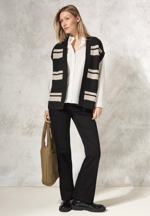 Black and cream striped knitted vest over a white shirt, paired with black flared pants and shiny black shoes, holding a tan tote bag.