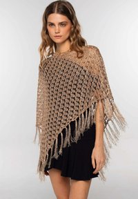 Poncho de ganchillo en beige claro con un diseño de tejido abierto, con detalles de flecos en los bordes. Lleva puesto sobre un vestido negro.