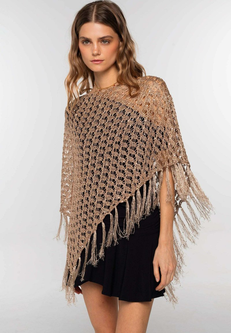 Poncho de ganchillo en beige claro con un diseño de tejido abierto, con detalles de flecos en los bordes. Lleva puesto sobre un vestido negro.