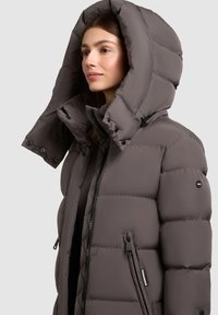 Žena v šedé zateplené zimní bundě s oversized kapucí na jednobarevném světlém pozadí.