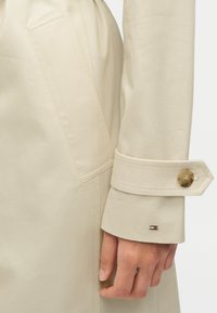 Μπεζ καμπαρντίνα με ομαλή υφή, διαθέτει πλαϊνή τσέπη, κουμπωτό μανίκι και μικρό λογότυπο κοντά στο μανίκι.