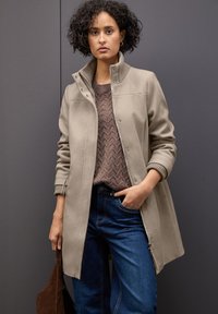 Beżowy płaszcz wełniany z wysokim kołnierzem, zamkiem błyskawicznym i napami; noszony nad brązowym swetrem o teksturze i niebieskimi dżinsami.