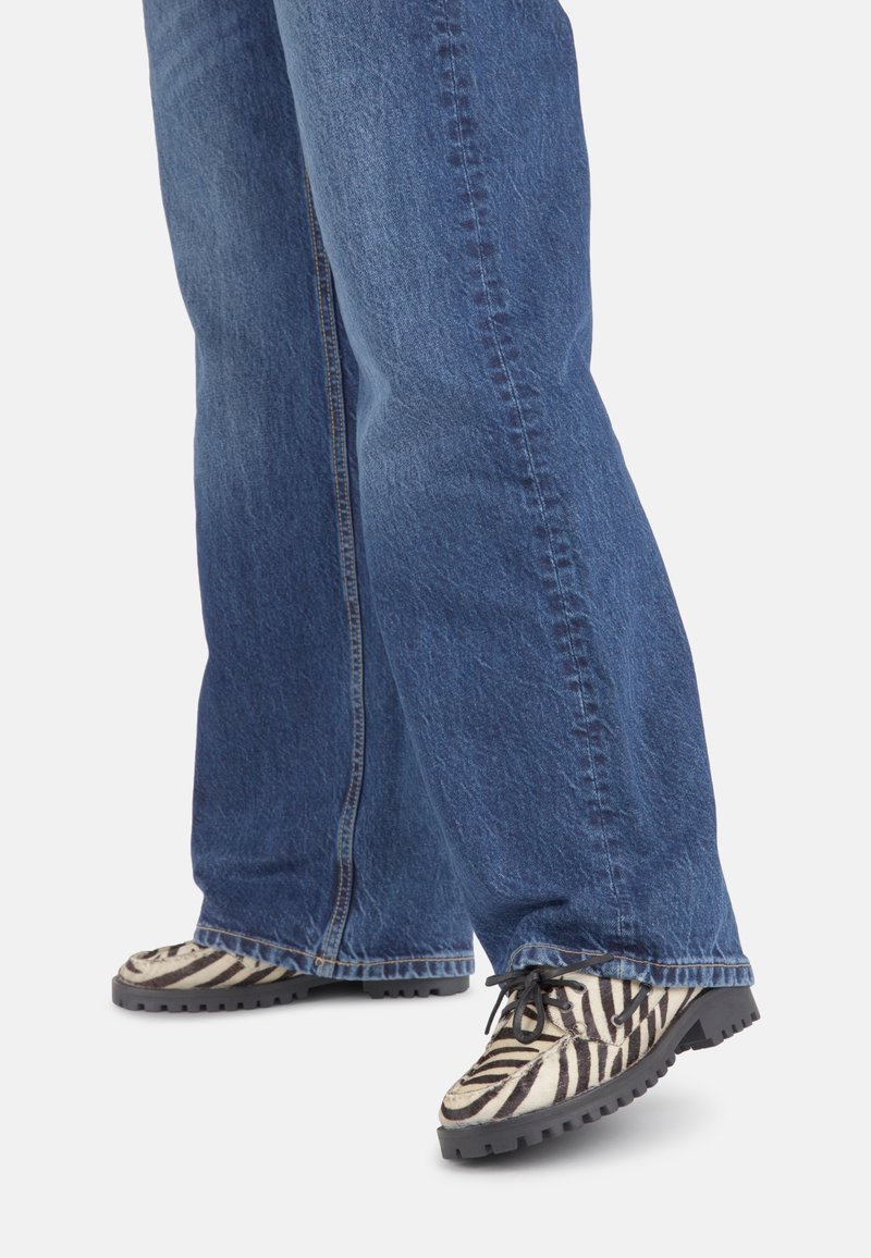 Des chaussures à motif zèbre avec des rayures noires et blanches, dotées d'une semelle robuste, associées à un jean en denim bleu à coupe ample.