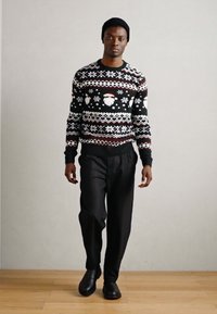 Pullover de malha em preto e branco com padrões de festa, combinado com calças largas pretas e botas pretas, usado por uma figura masculina em pé.