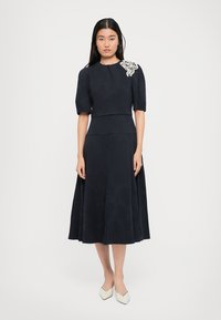 Marineblauwe jurk met korte pofmouwen en een getailleerde bodice. Heeft een gestructureerde stof en witte borduursel op de linkerschouder.