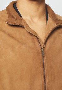 Béžová sukně z veluru s žebrovaným límcem, zapínáním na zip a hladkým povrchem, který zdůrazňuje čistý a minimalistický design.