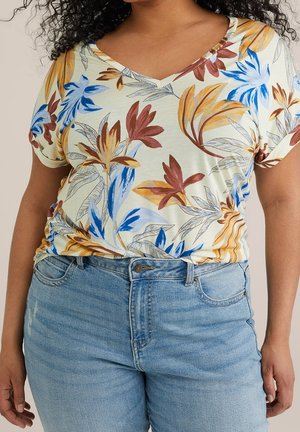 Kvinde iført en V-hals kortærmet skjorte med flerfarvet blomsterprint, stoppet ned i lyseblå høj-taljede jeans.