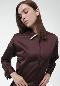 Camisa de satén marrón con cuello y acento dorado, que presenta mangas largas y un ajuste estructurado. Textura suave y diseño minimalista.