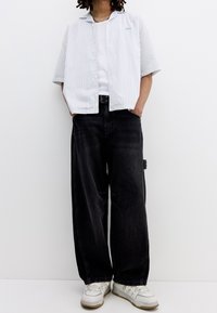 Košile s krátkým rukávem, bílá s proužky, zapínaná na knoflíky s límečkem, v kombinaci s širokými černými džínovými kalhotami a bílými teniskami. Texturovaná látka s viditelným prošitím.