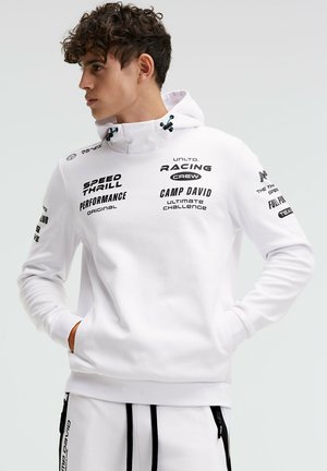 Young man wearing white hooded sweatshirt and matching pants with black text logos, hands in pockets, looking to his left.