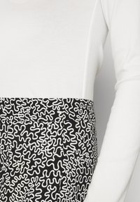 Gros plan d'une personne portant un haut blanc à manches longues rentré dans un pantalon noir avec un motif abstrait blanc en zigzag.