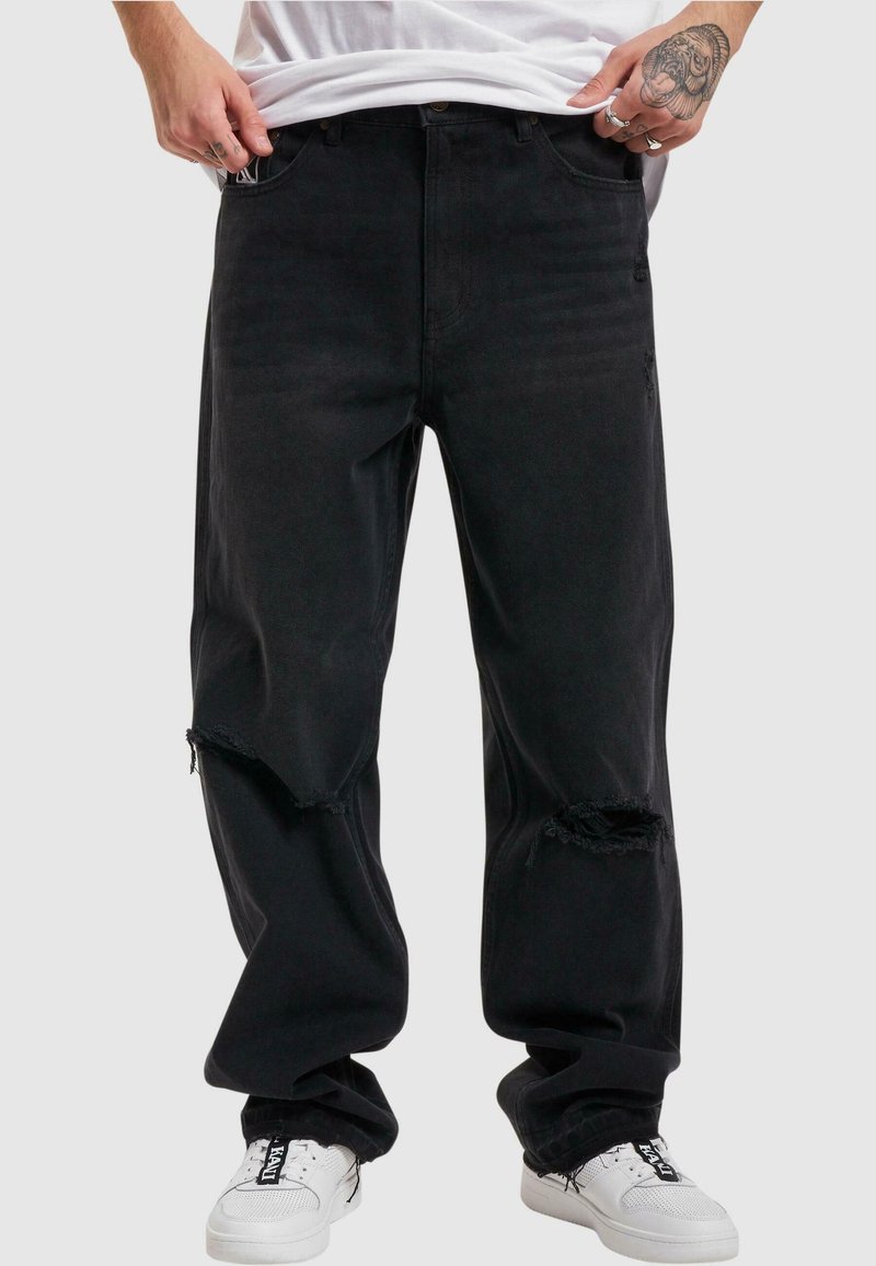 Jeans de mezclilla negra con un corte relajado, rodillas desgastadas con bordes deshilachados y diseño de cinco bolsillos. Llevados con zapatillas blancas.