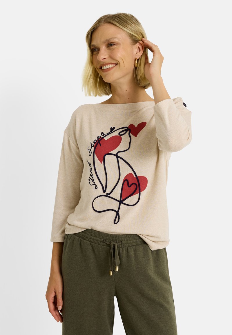 Pull léger beige avec un design graphique présentant des lignes entrelacées et des cœurs rouges. Texture douce, avec des manches trois-quarts et une coupe décontractée.