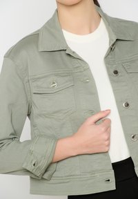 Zbliżenie na osobę w jasnoniebieskiej dżinsowej kurtce z srebrnymi guzikami, noszącą białą bluzkę, trzymającą przód kurtki prawą ręką.