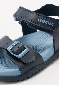 Sandal com biqueira aberta, feita de material sintético azul marinho, com sola de borracha azul. Apresenta uma tira com um fecho azul e uma palmilha texturizada.