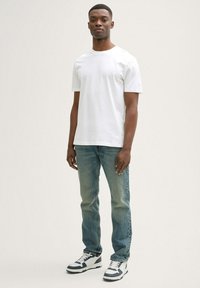 Biała bawełniana koszulka z krótkim rękawem, połączona z jasnoniebieskimi dżinsami i czarno-białymi sneakersami, stojąca na jasnym tle.