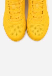 Baskets de sport jaunes avec une tige en caoutchouc lisse, un design perforé et des lacets plats. Bout arrondi et semelle texturée pour une meilleure adhérence.