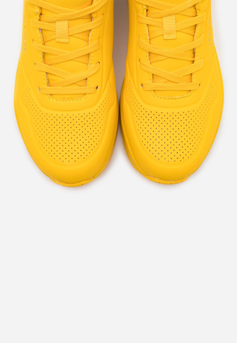 Zapatillas deportivas amarillas con un upper de goma suave, diseño perforado y cordones planos. Punta redondeada y suela texturizada para un mejor agarre.