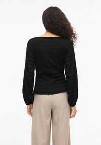 Czarna bluzka z długim rękawem z teksturowanej tkaniny, z dekoltem w łódkę i marszczonymi mankietami, zestawiona z jasnobeżowymi spodniami o szerokich nogawkach. Ząbkowany detal na dole.