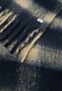 Plava i bež karirana vuna deka s rastrganim rubovima i oznakom branda. Tekstilirana tkanina sadrži različite nijanse plave boje.
