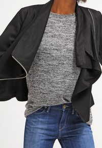 Chaqueta ligera negra con delantero asimétrico y cremallera plateada, llevada sobre un suéter gris texturizado. Combinada con jeans azules.