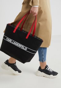 Personne portant un trench beige et un jean bleu tient un sac cabas noir Karl Lagerfeld avec des anses rouges, portant des baskets noires et blanches.