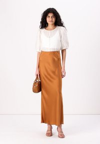 Blusa bianca, con texture, maniche a palloncino e chiusura con bottoni, abbinata a una gonna lunga in satin arancio-marrone liscia. Borsa beige e sandali.