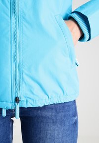 Lichtblauwe jas van gladde stof, met een ritssluiting en geribde boorden, voorzien van zijzakken en een zoom met trekkoord.