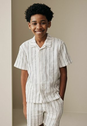 Mosolygó fiú rövid, göndör hajjal, fehér rövid ujjú, gombos ingben és hozzáillő csíkos rövidnadrágban, egyik kezét a zsebébe dugva áll.